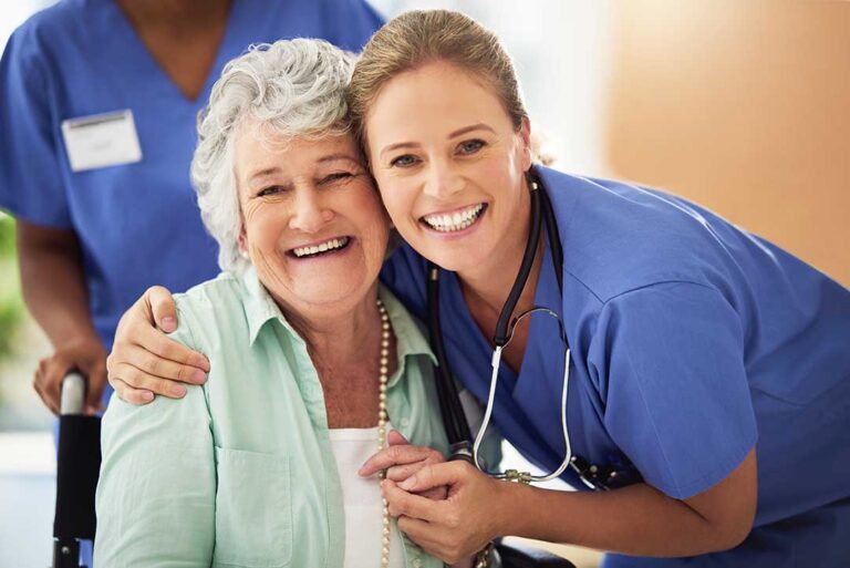 Middle-aged adult holding hands and talking with an elderly parent in an assisted living room.