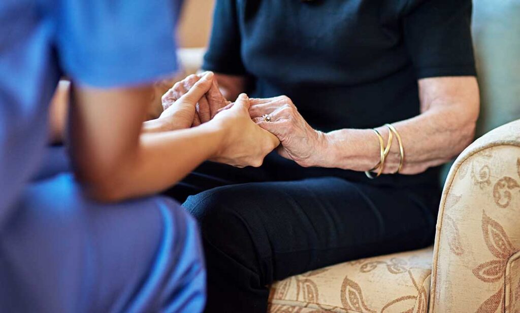 A senior resident receives a blood pressure check from a caregiver in a home-like living space.